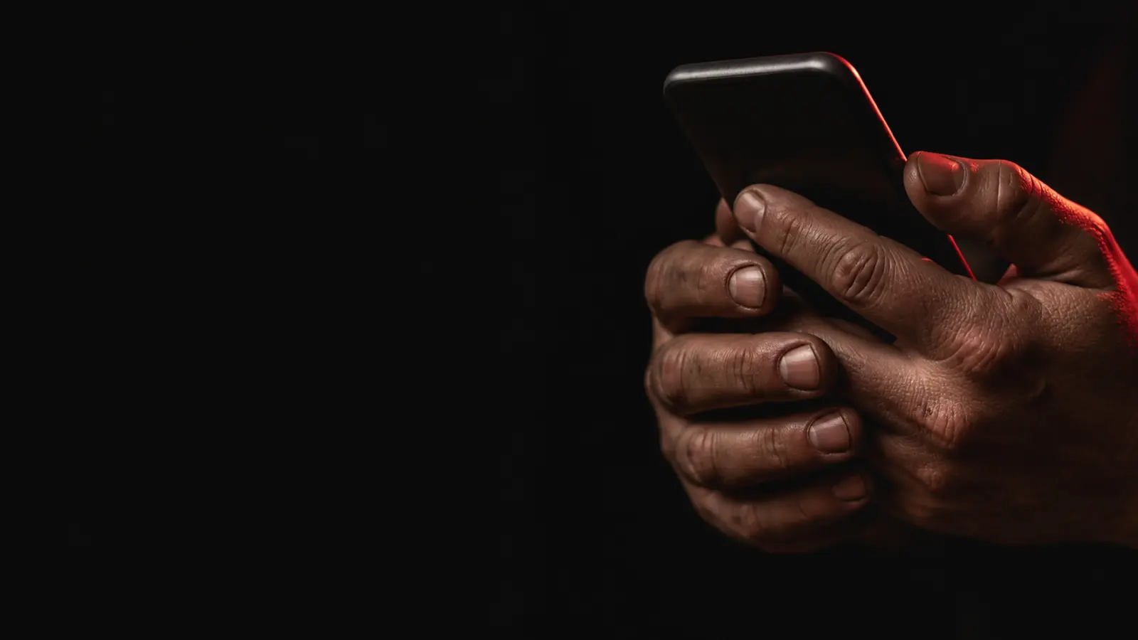 Deux mains d'artisan tiennent un téléphone dans la pénombre d'un atelier ; une lueur rouge court sur le bord de l'appareil.